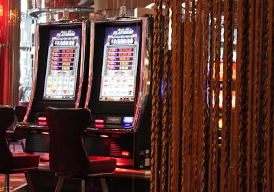 A pair of casino slot machines, each with detailed screens displaying various game outcomes and jackpot amounts. The chairs in front of the slot machines are dark and cushioned for comfort. The background features a hanging curtain made of golden ropes and other casino decorations are visible.