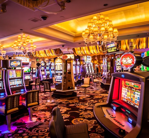 A vibrant casino interior filled with rows of colorful slot machines, each illuminated with bright lights and displays. The ceiling is adorned with elegant chandeliers and golden drapes, creating a luxurious atmosphere. The patterned carpet adds to the elaborate design of the space.