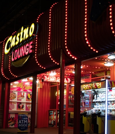 A brightly lit casino lounge entrance adorned with neon lights. The exterior features colorful signage including the words 'Casino' and 'Lounge' in vivid colors, with additional lights framing the building. Inside, a glimpse of a claw machine filled with plush toys and arcade games is visible. The area exudes an inviting yet bustling atmosphere typical of entertainment venues.