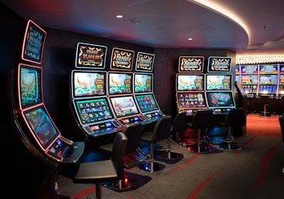 A row of brightly lit slot machines in a casino setting, each with colorful displays and various game themes. The machines are lined up against a dark wall and are unoccupied, with cushioned chairs placed in front of them. The area is dimly lit except for the glow from the screens, creating a vibrant and engaging atmosphere.